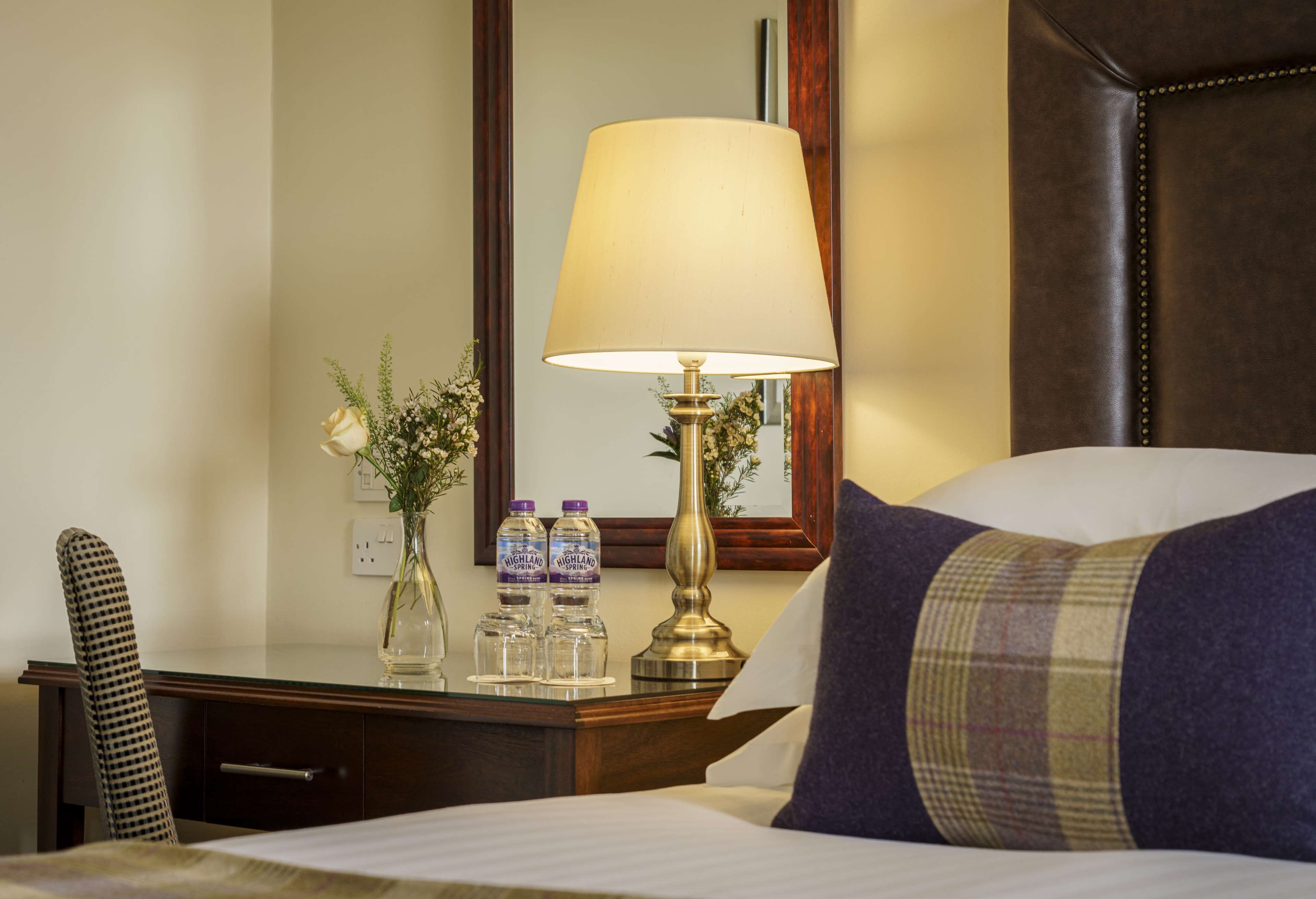Une chambre d’hôtel avec un lit, une table de chevet avec une lampe, un miroir, un vase avec des fleurs et deux bouteilles d’eau.