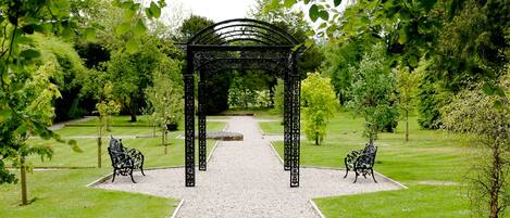 A garden pathway leading to a black wrought iron archway with benches on either side.