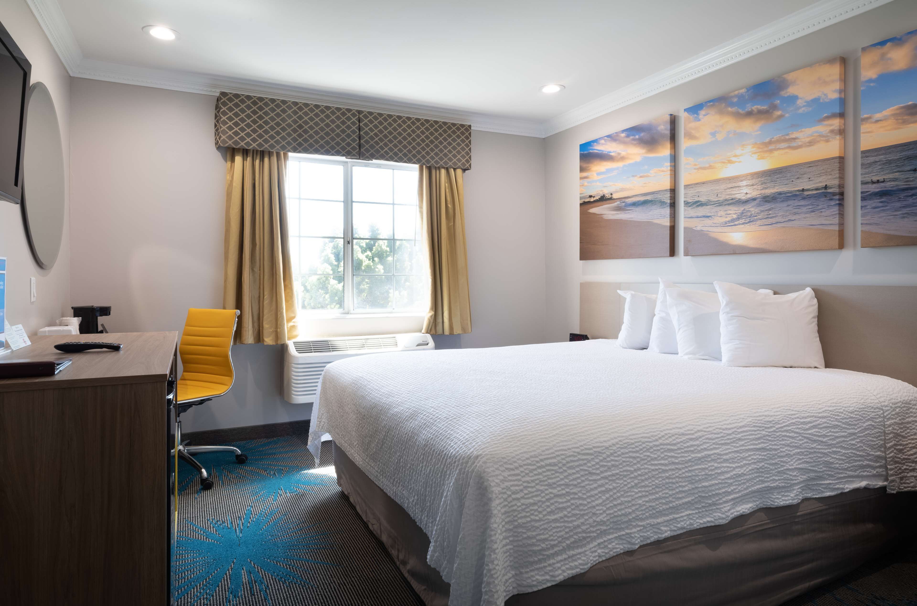 Habitación de hotel con cama, escritorio, silla, ventana con cortinas y una pintura de un atardecer sobre el océano.