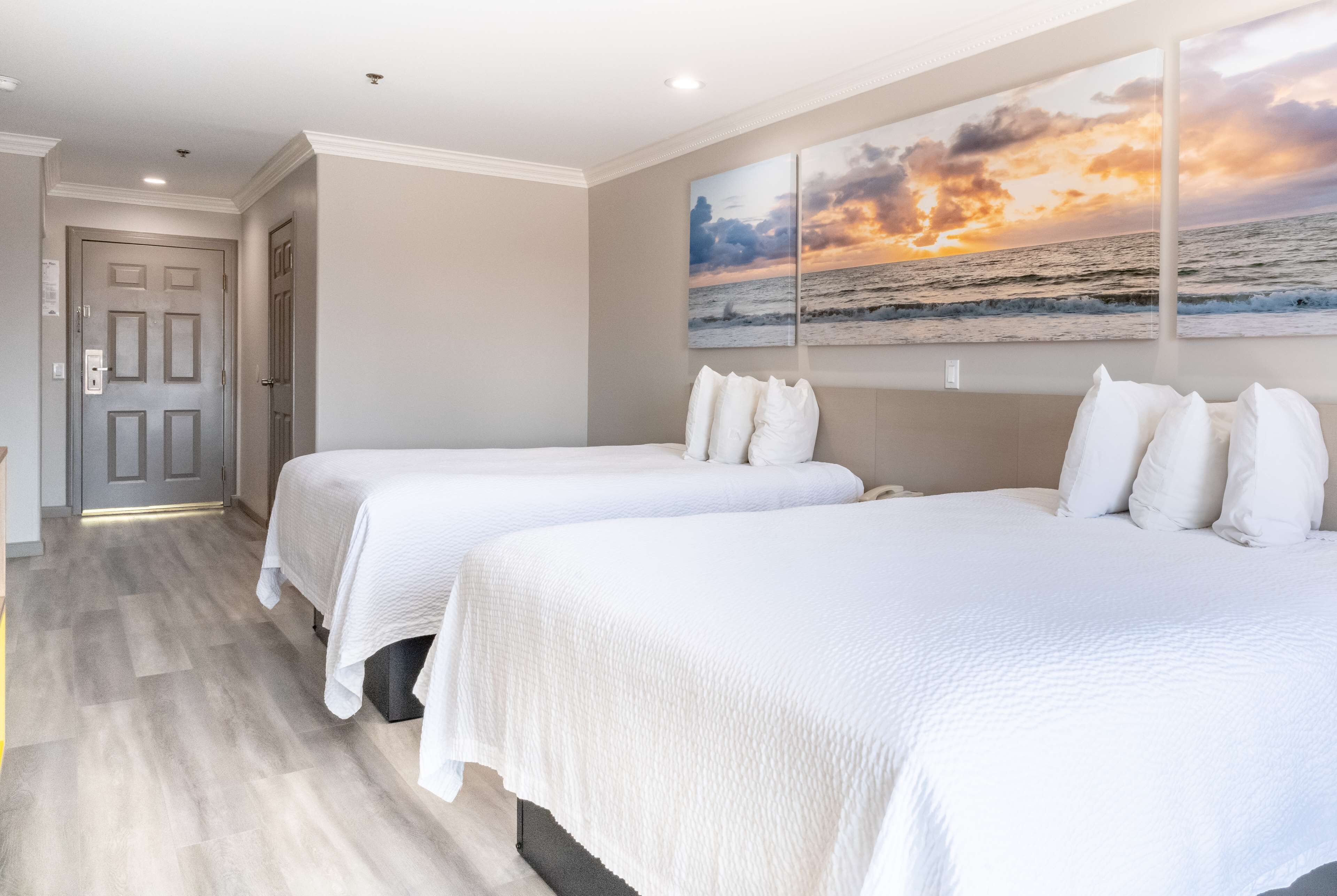 Habitación de hotel con dos camas, una puerta y un cuadro con atardecer sobre el océano.