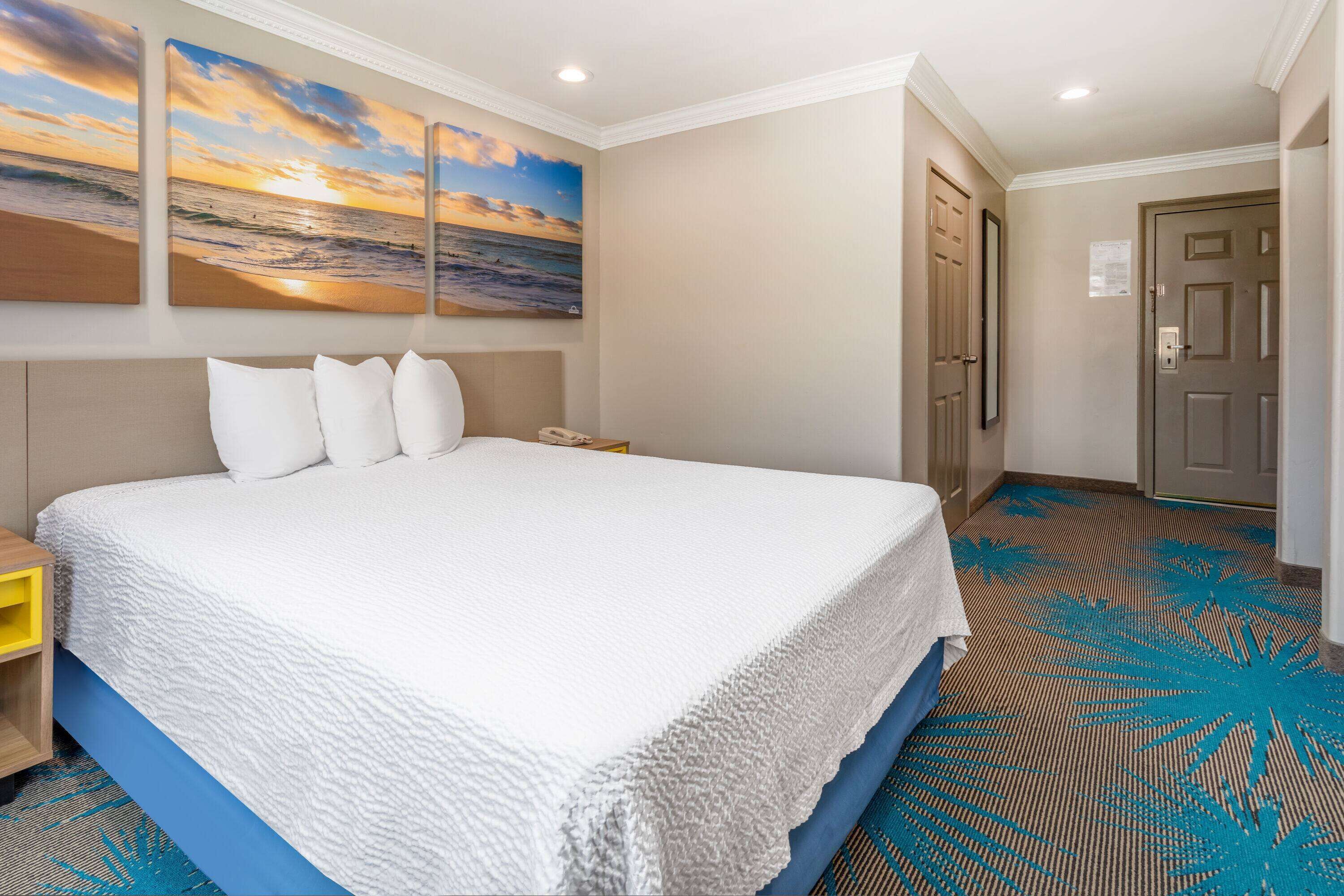 Habitación de hotel con una cama grande, una mesita de noche, un cuadro de un atardecer sobre el océano y una puerta a la derecha.