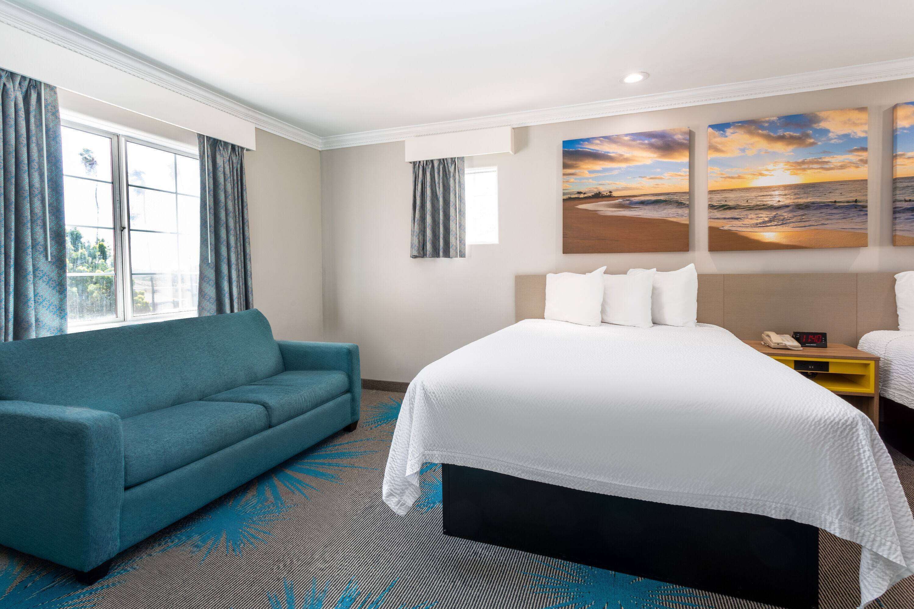 Habitación de hotel con cama, sofá, mesita de noche y un cuadro de una puesta de sol sobre el océano.