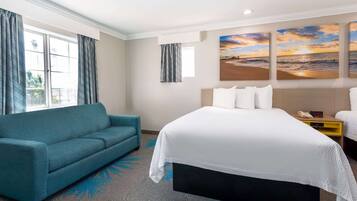 Habitación de hotel con cama, sofá, mesita de noche y un cuadro de una puesta de sol sobre el océano.