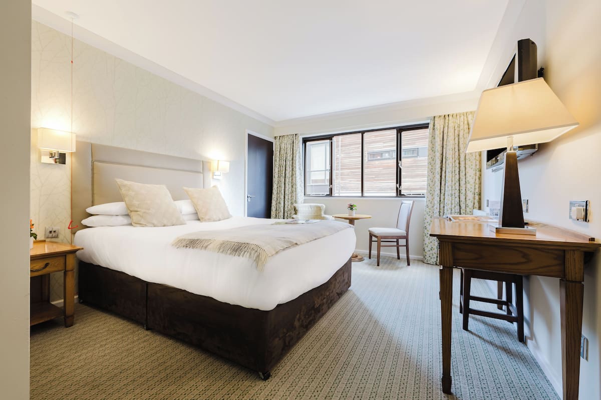 A hotel room with a large bed, a desk, a chair, a lamp, and a window with curtains.