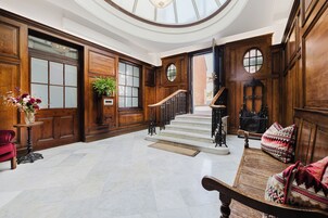 Un amplio vestíbulo con suelo de mármol, paneles de madera, una escalera y un banco con cojines estampados.