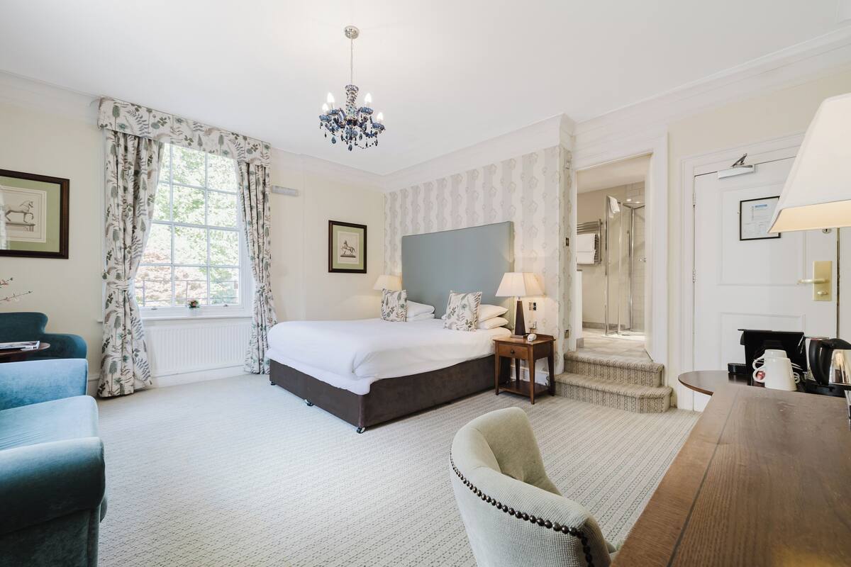 A hotel room with a bed, a chair, a table, a lamp, a chandelier, and a window with curtains.