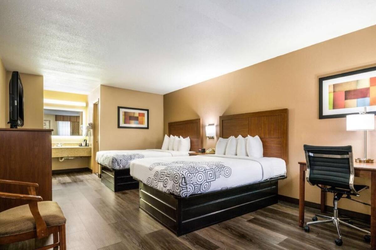 A hotel room with two beds, a desk, a chair, a TV, and a bathroom visible in the background.