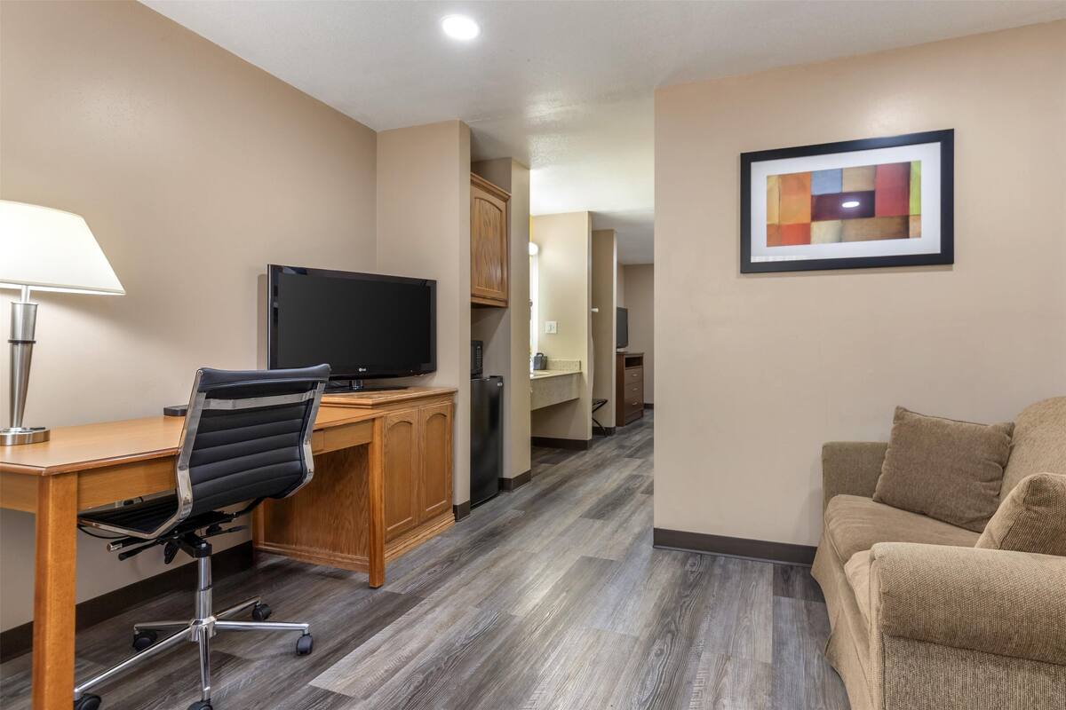 A hotel room with a wooden desk, a black office chair, a television, a beige sofa, and a framed abstract painting on the wall.
