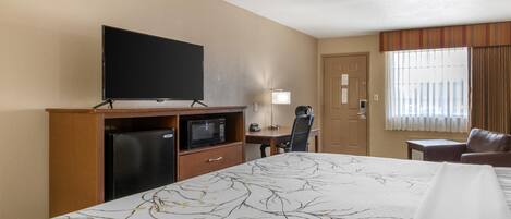 A hotel room with a bed, a desk with a computer, a television, a refrigerator, and a window with curtains.