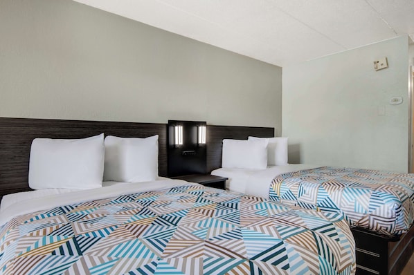 Habitación de hotel con dos camas, cada una con una colcha estampada y almohadas blancas. Hay una mesita de noche con lámpara entre las camas, y la habitación presenta un diseño sencillo y limpio con cabecero de madera y pared de color claro.