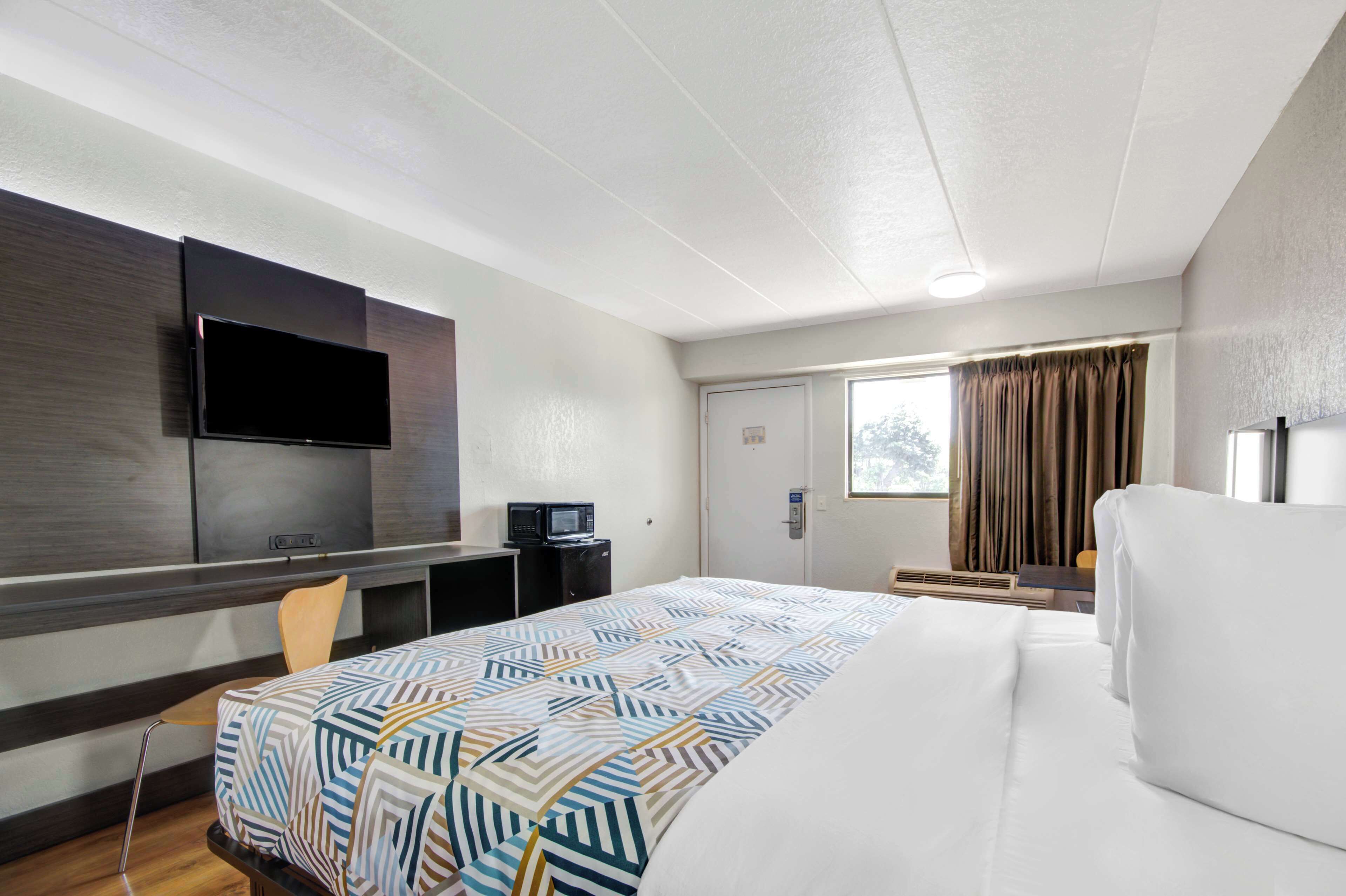 A hotel room with a bed, a desk with a flat-screen TV, a microwave, and a window with curtains.