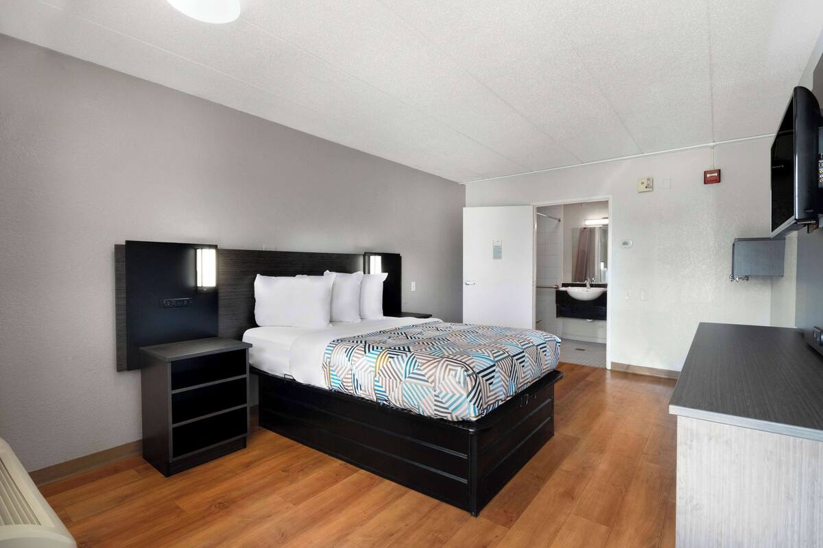 A modern bedroom with a black bed frame, a bedside table, a built-in TV, and a bathroom visible through an open door.