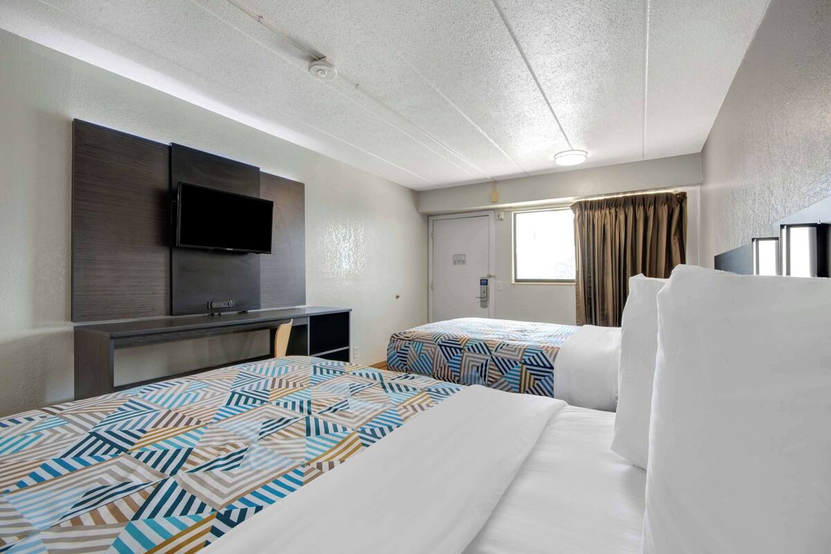 A hotel room with two beds, a desk, a flat-screen TV, and a window with curtains.