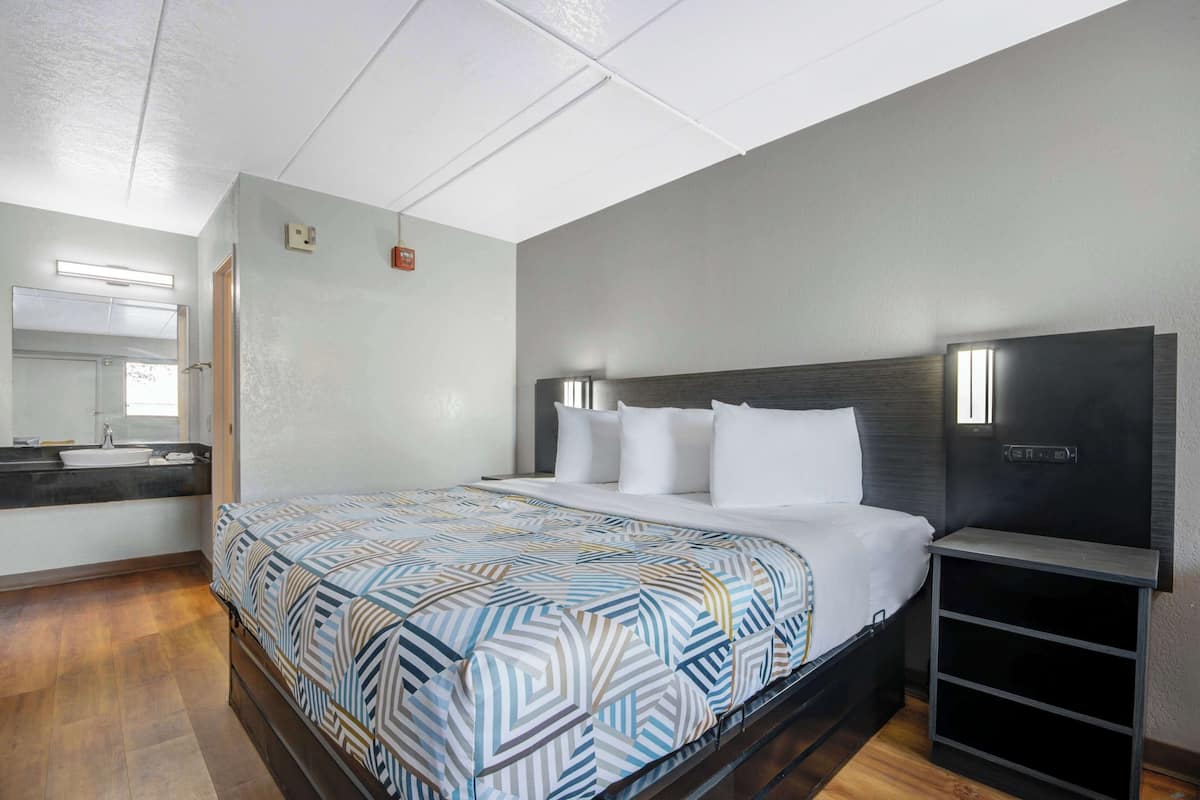 A hotel room with a large bed, a bedside table, a built-in TV, and a bathroom with a sink and mirror.