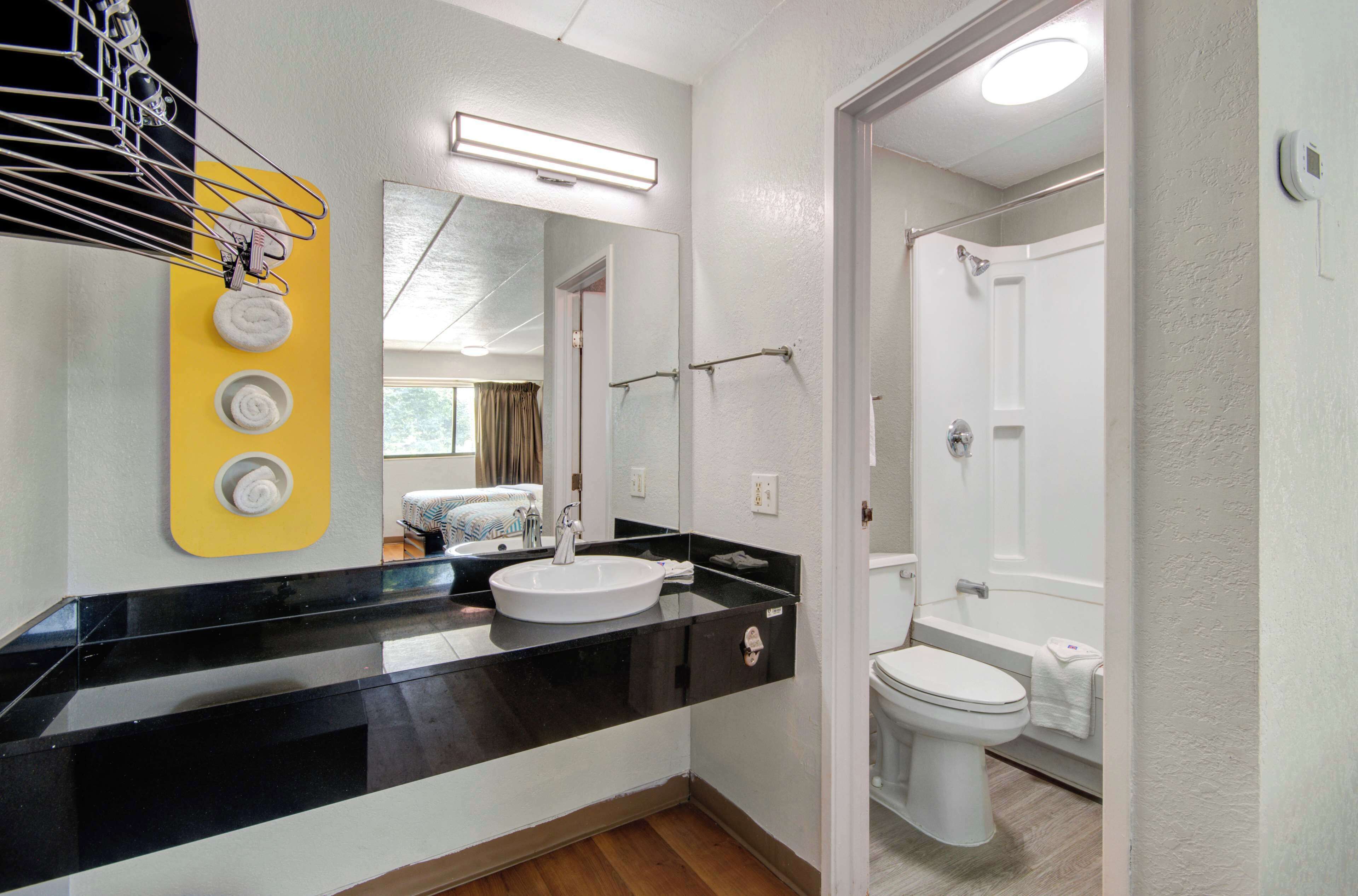 A modern bathroom with a black countertop, white sink, and a mirror above it. There is a shower area visible in the background.