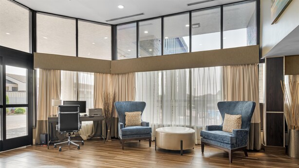 A modern hotel lobby with large windows, a wooden floor, two blue armchairs, a round ottoman, and a desk with a computer. - Best Western Center Inn (Virginia Beach)