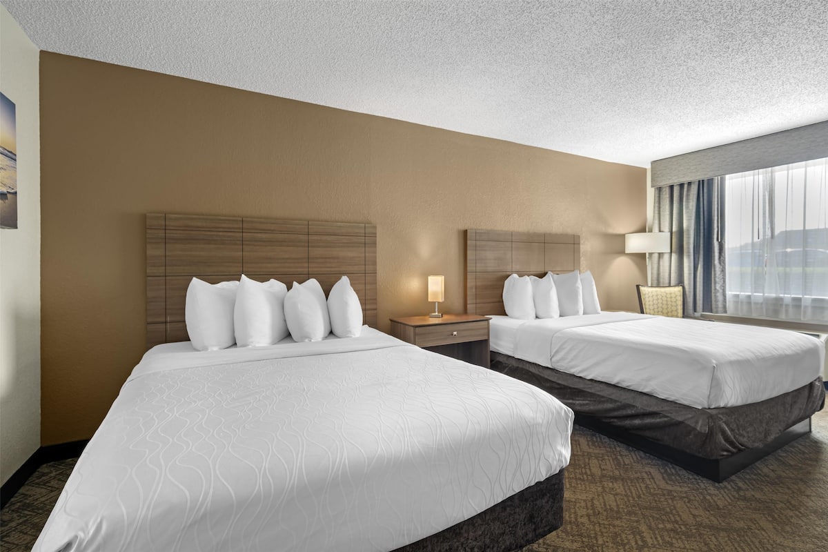 A hotel room with two beds, a nightstand, a lamp, and a window with curtains.