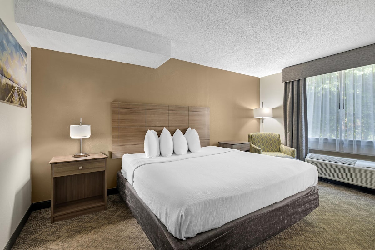 A hotel room with a large bed, a wooden headboard, a nightstand with a lamp, a chair, and a large window with curtains.