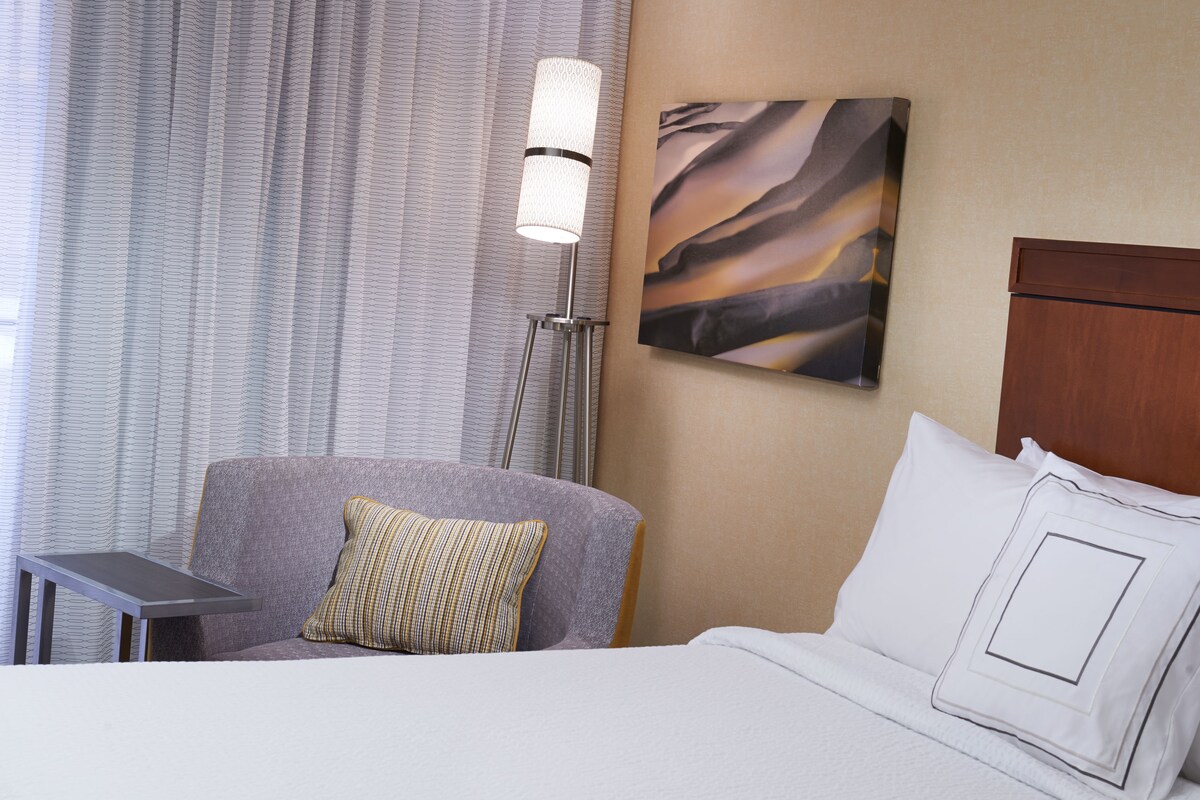 A hotel room with a bed, a chair, a floor lamp, and a painting on the wall.