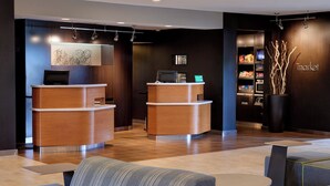 A modern reception area with two wooden reception desks, a seating area with sofas and a round table, and a wall-mounted display shelf with various items. - Courtyard by Marriott Detroit Dearborn (Dearborn)