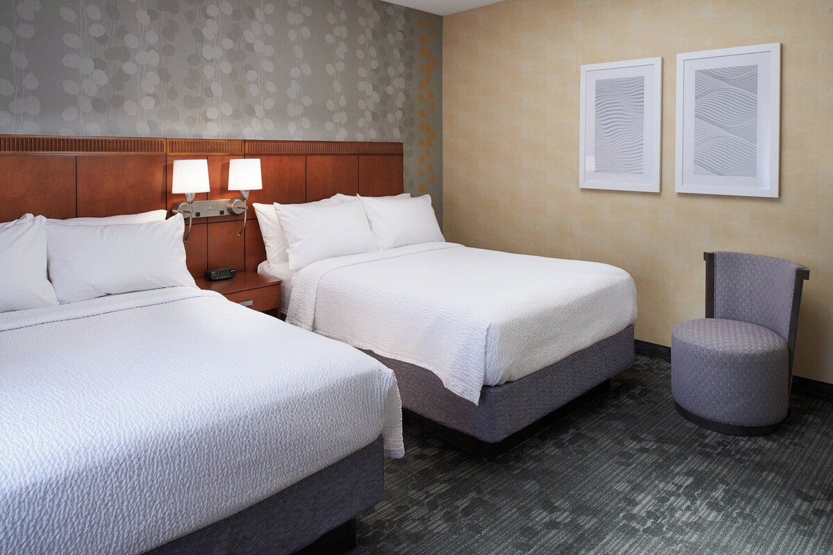 A hotel room with two beds, a nightstand with lamps, a chair, and two framed artworks on the wall.