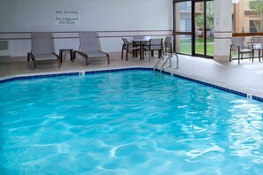 Piscina interna com espreguiçadeiras e uma mesa, um aviso indicando 'NÃO MERGULHAR Sem salva-vidas' e vista para o exterior.