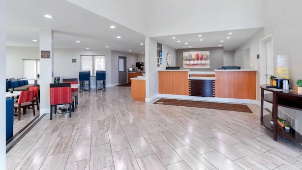 A spacious hotel lobby with a reception desk, a dining area with red upholstered chairs, and a kitchen area.