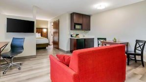 A hotel room with a kitchenette, a TV mounted on the wall, a bed, and a red armchair.