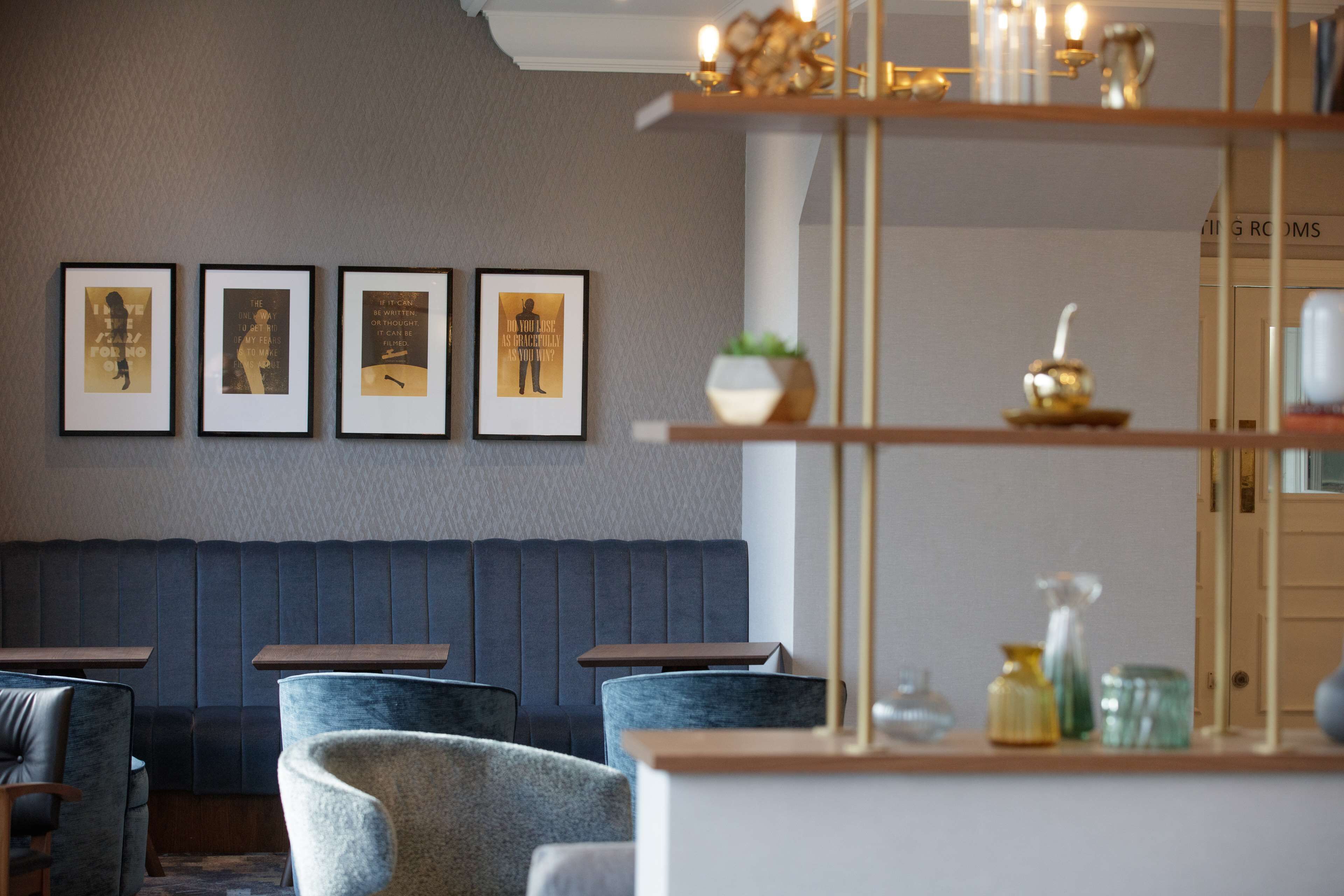 Un interior moderno con una zona de estar que incluye sillas acolchadas y un banco de color oscuro. En la pared hay obras de arte enmarcadas y una estantería con objetos decorativos.