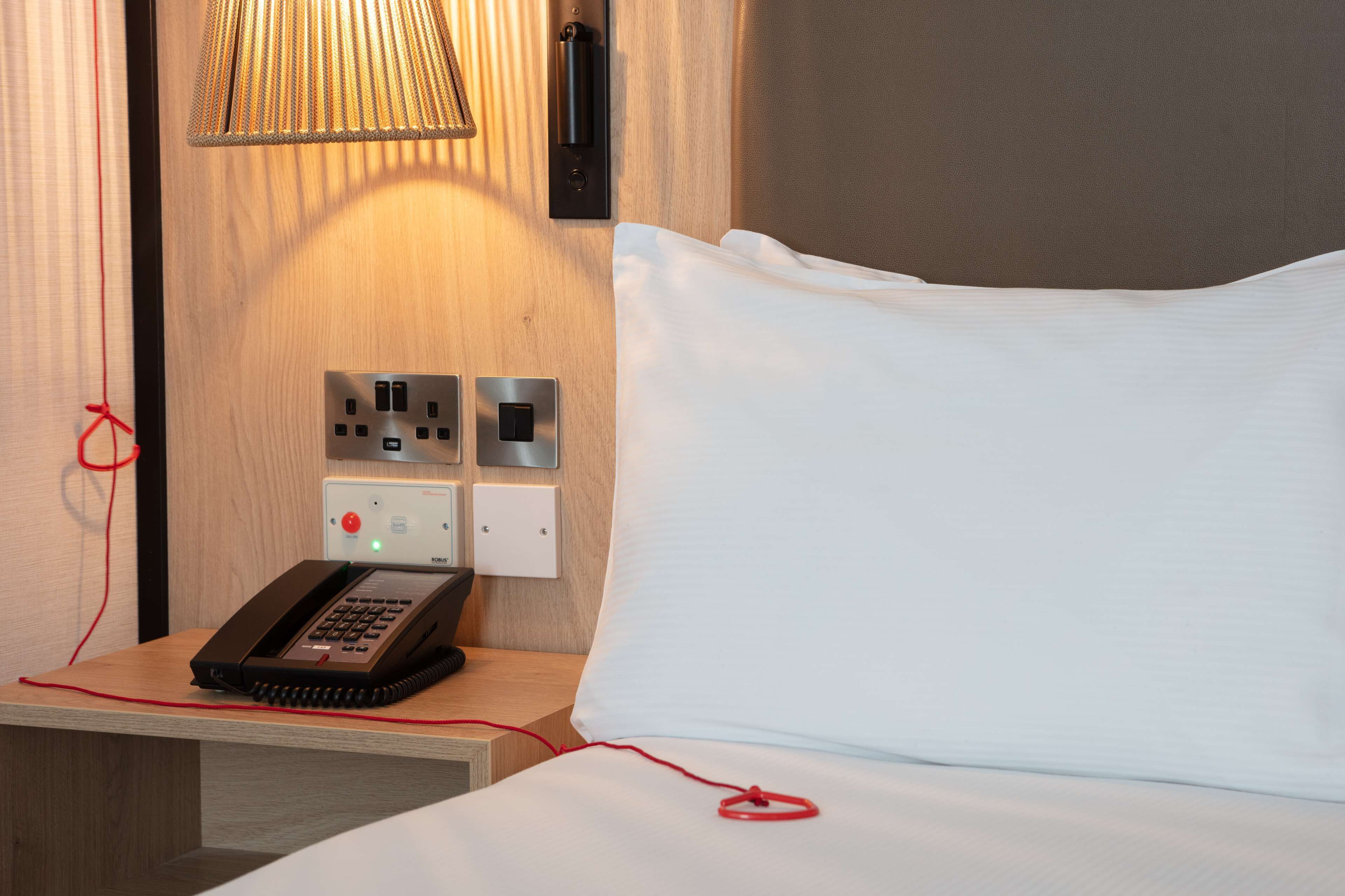 Habitación de hotel con cama, teléfono en la mesita de noche, lámpara y enchufes en la pared.