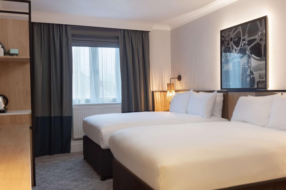 A hotel room with two beds, a large window with curtains, a wall-mounted lamp, and a framed artwork.