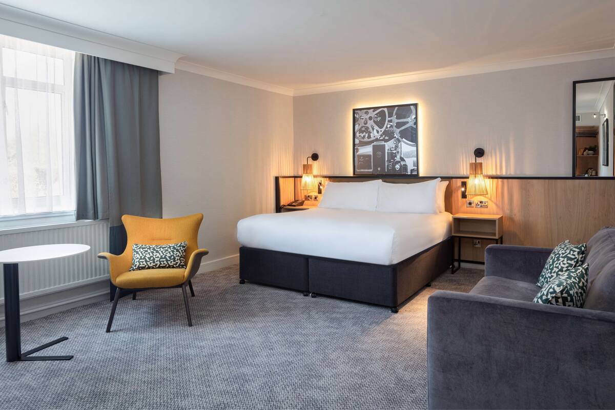 A hotel room with a bed, a chair, a sofa, a small table, and a window with curtains.