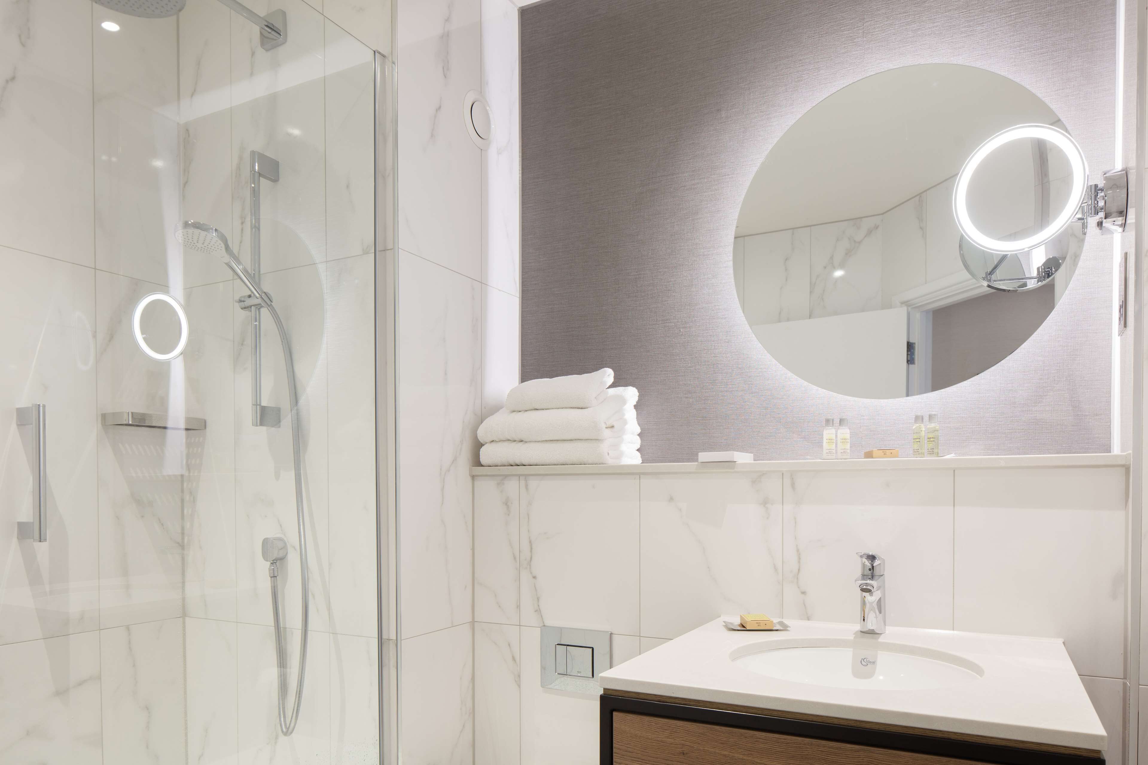 Een moderne badkamer met een douchecabine van glas, een ronde spiegel met verlichting en een witte wastafel.