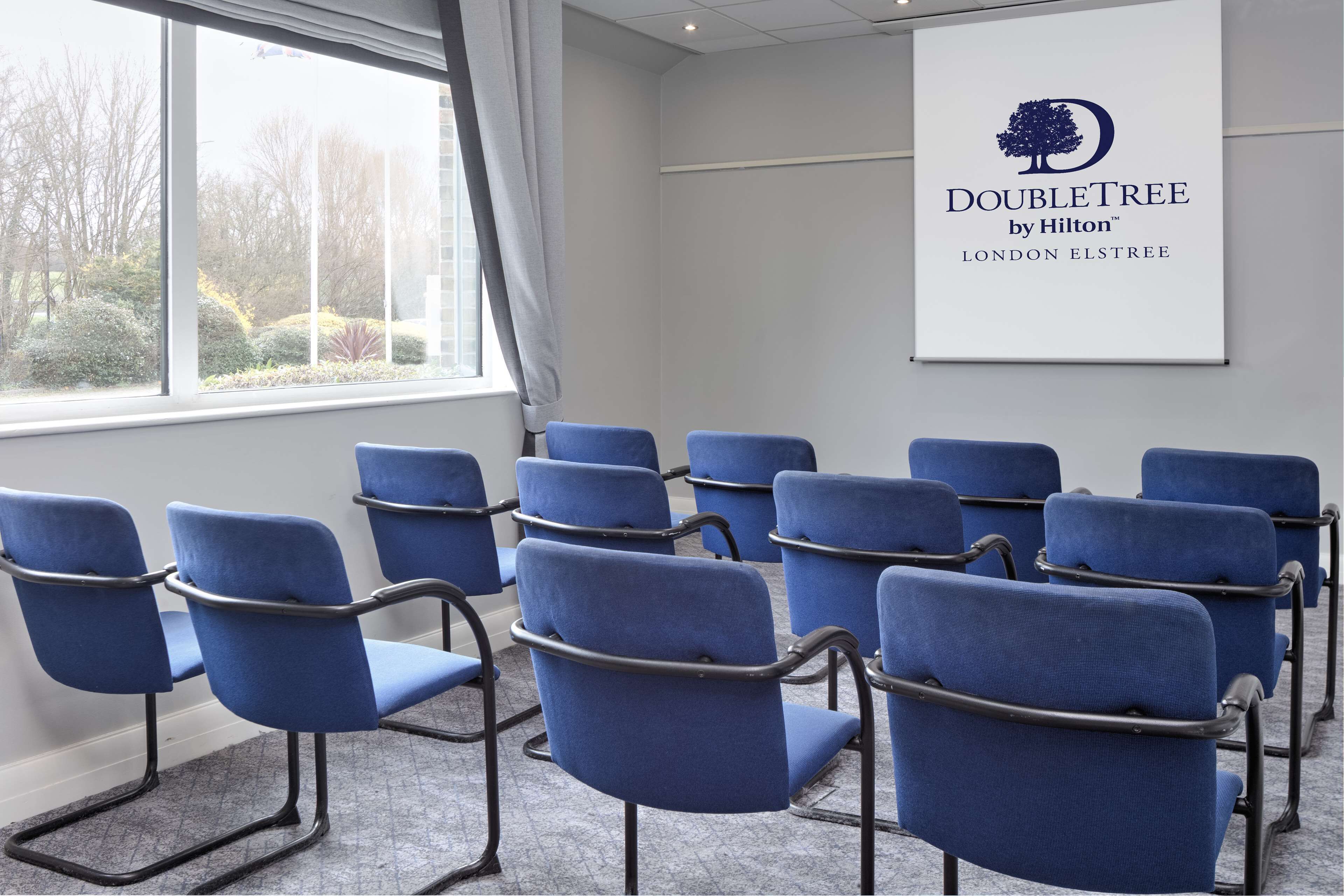 A conference room with blue chairs arranged in rows, a large window with a view of trees, and a wall-mounted sign for DoubleTree by Hilton London Elstree.