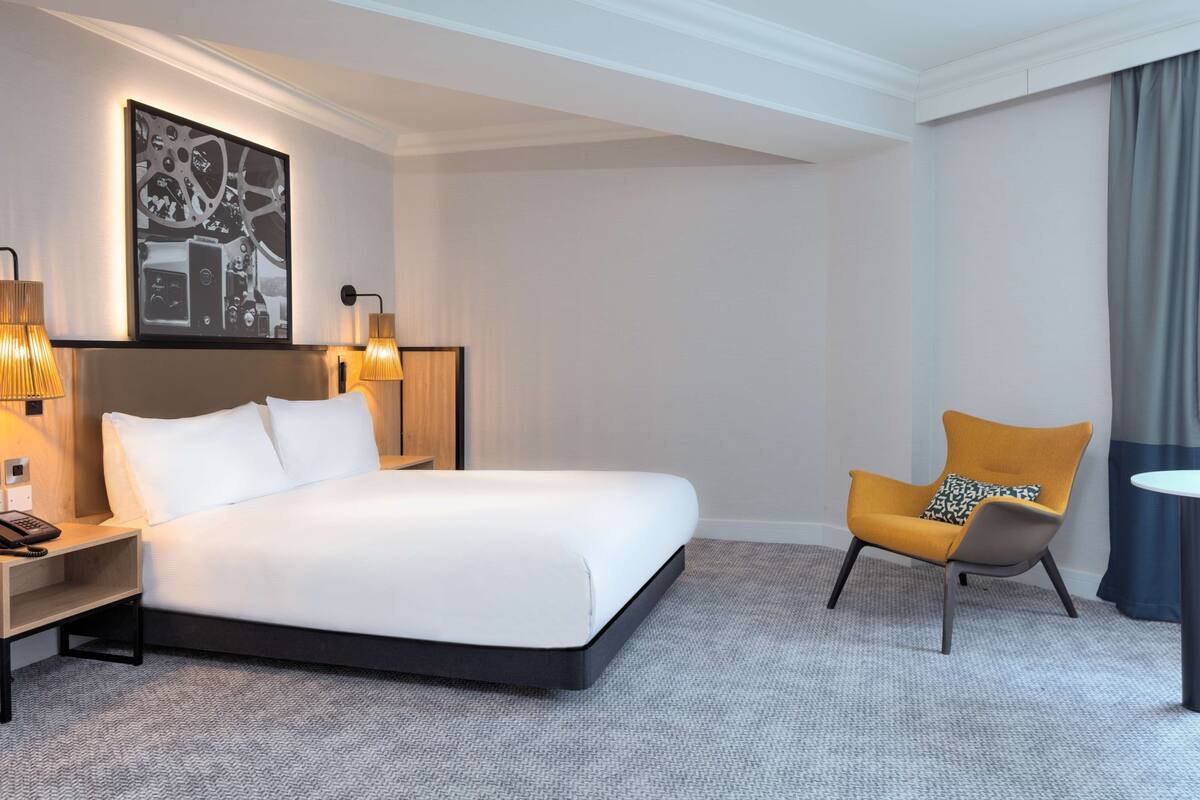 A modern hotel room with a large bed, a side table with a lamp, a chair, and a painting on the wall.
