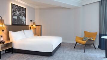 A modern hotel room with a large bed, a side table with a lamp, a chair, and a painting on the wall.