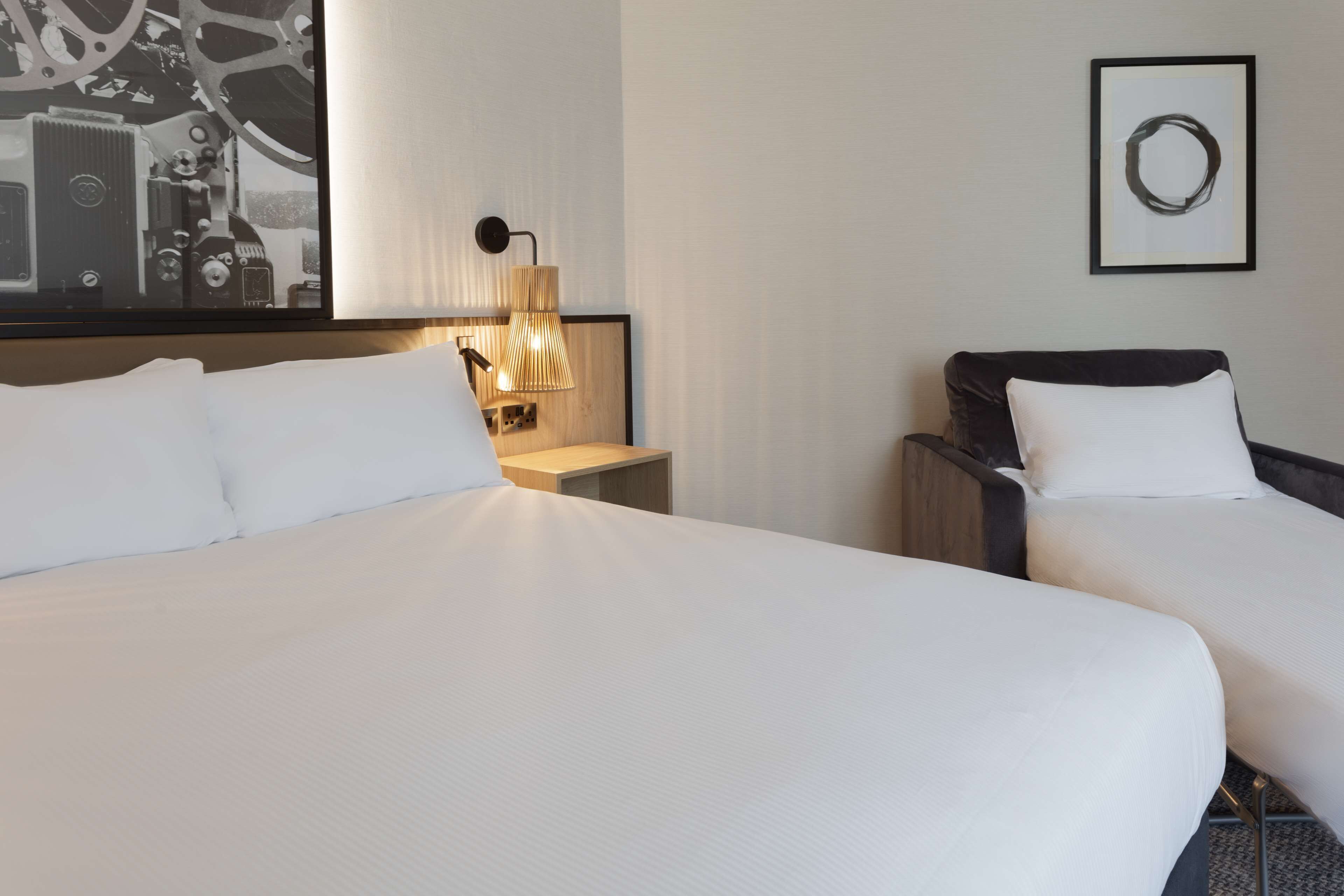 Quarto de hotel com duas camas, uma pintura abstrata grande, uma luminária fixada na parede e uma moldura com uma imagem.