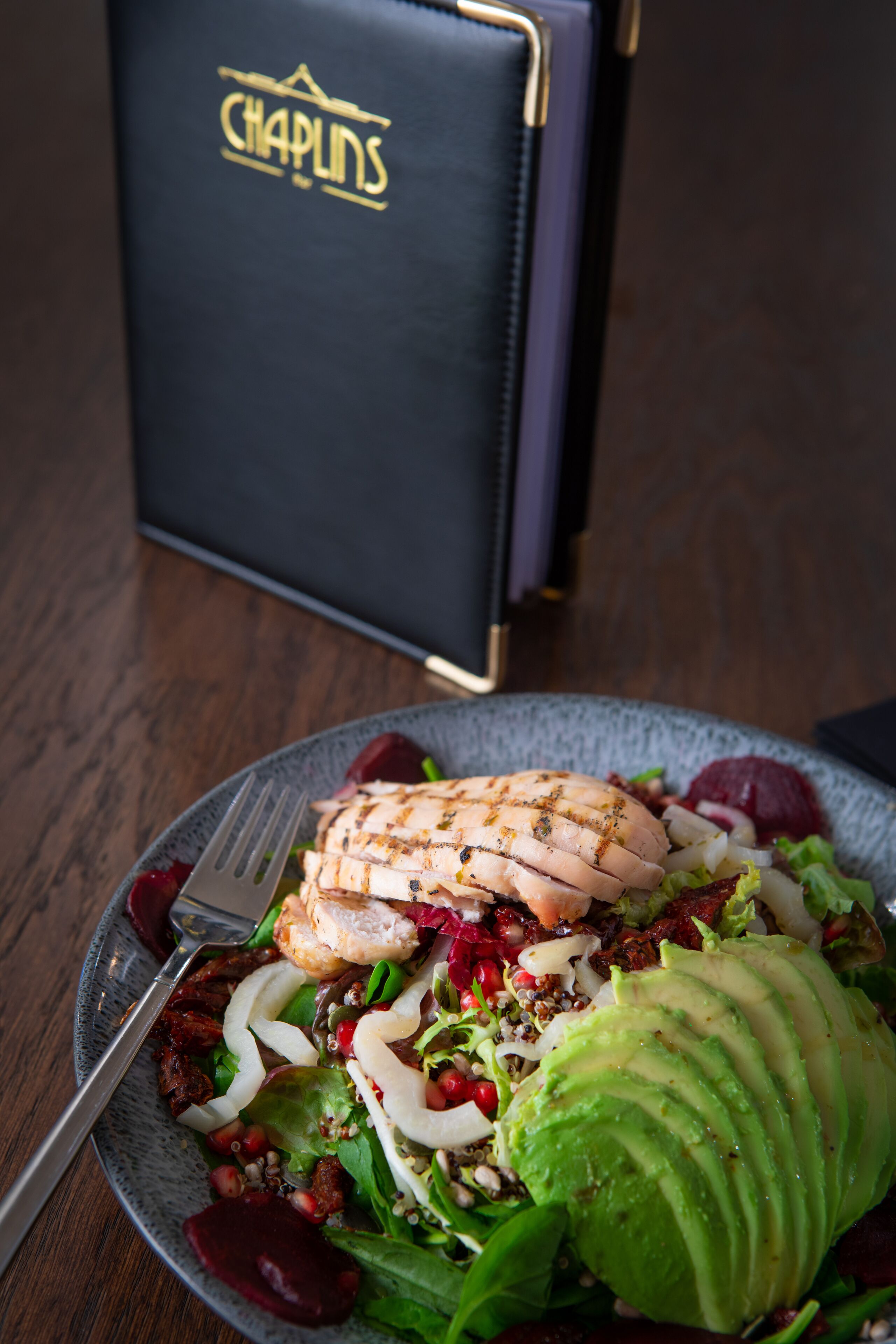 Ein Teller mit gegrilltem Hähnchen, Avocado und Salat auf einem Tisch, im Hintergrund eine dunkelblaue Speisekarte.