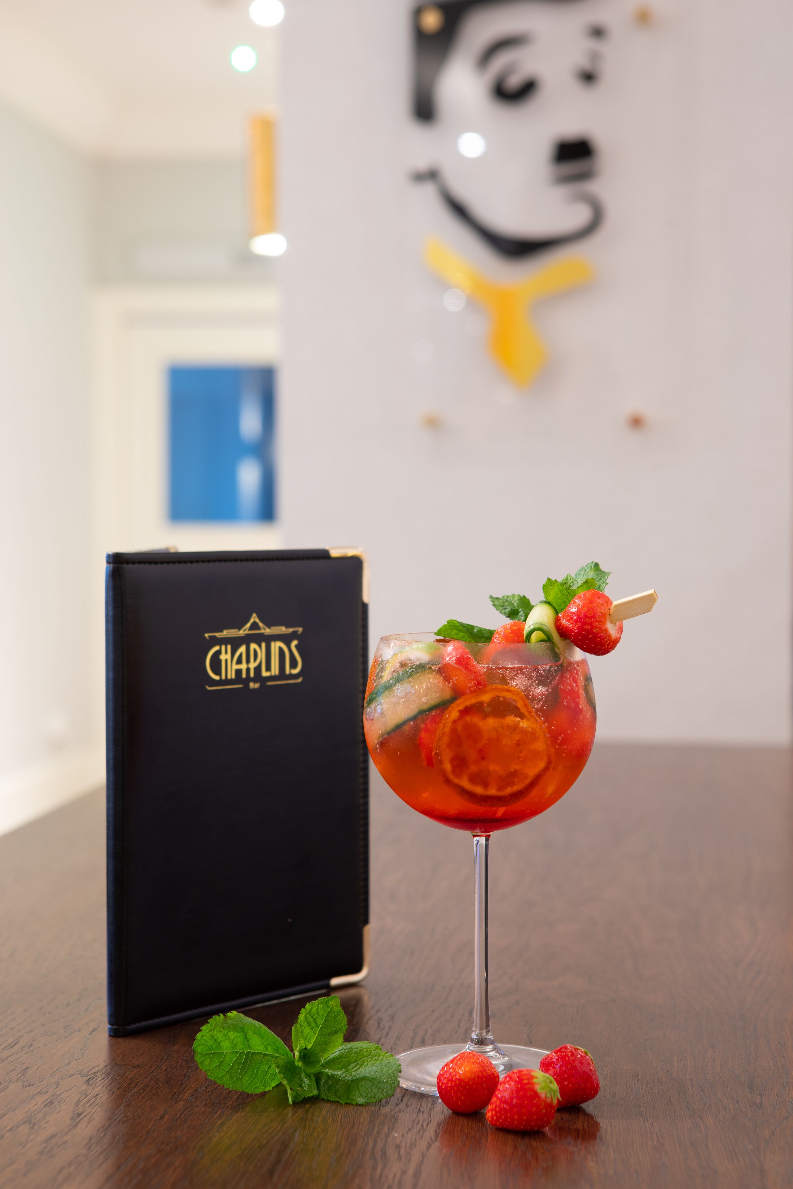 Una copa de cóctel con fresas y menta sobre una mesa de madera, una caja negra con el logotipo 'Chaplins' y un fondo desenfocado con una obra de arte en la pared.