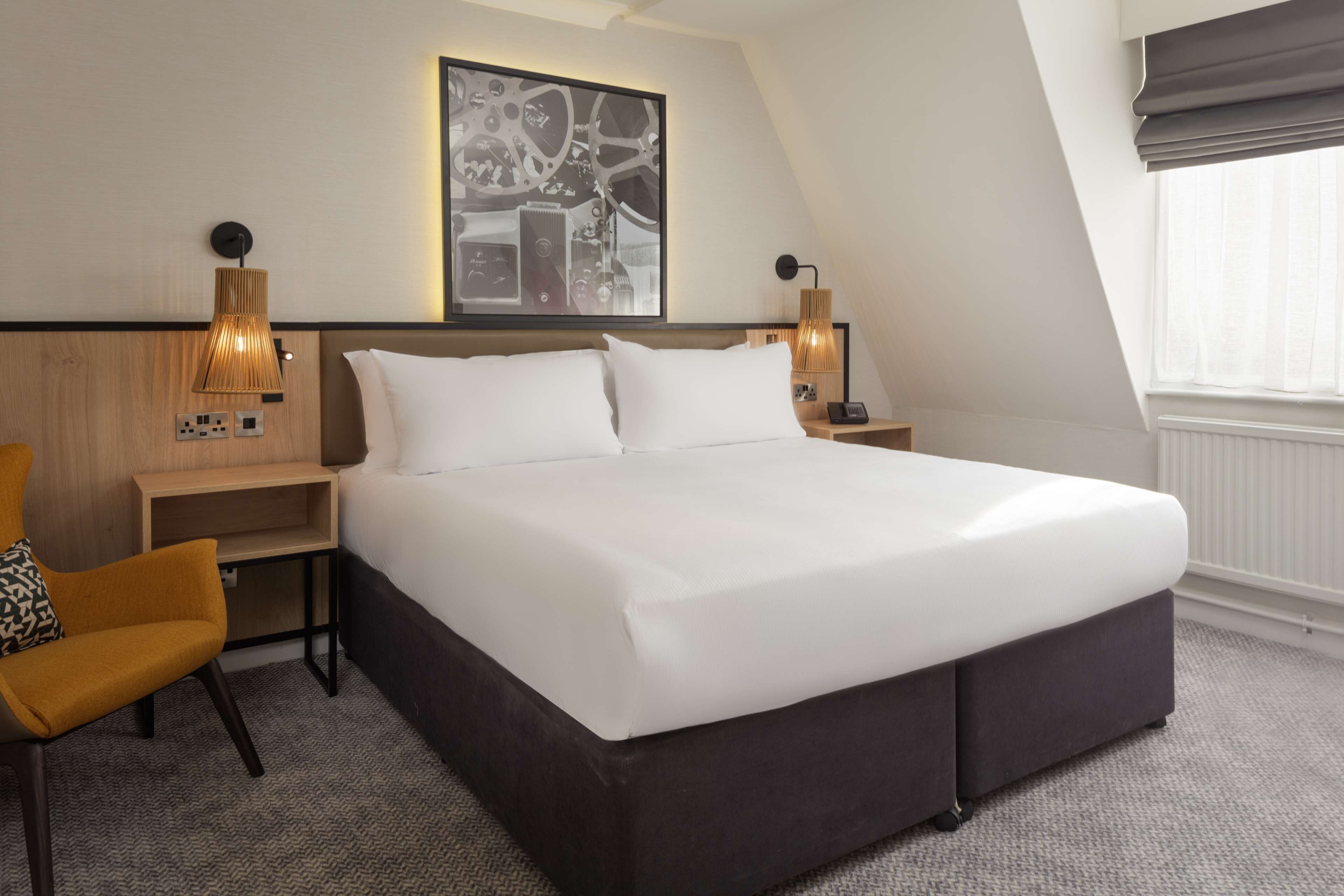 Une chambre d’hôtel moderne avec un grand lit, deux tables de chevet avec des lampes, une œuvre d’art accrochée au mur et une fenêtre avec des stores.