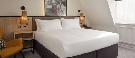 Une chambre d’hôtel moderne avec un grand lit, deux tables de chevet avec des lampes, une œuvre d’art accrochée au mur et une fenêtre avec des stores.