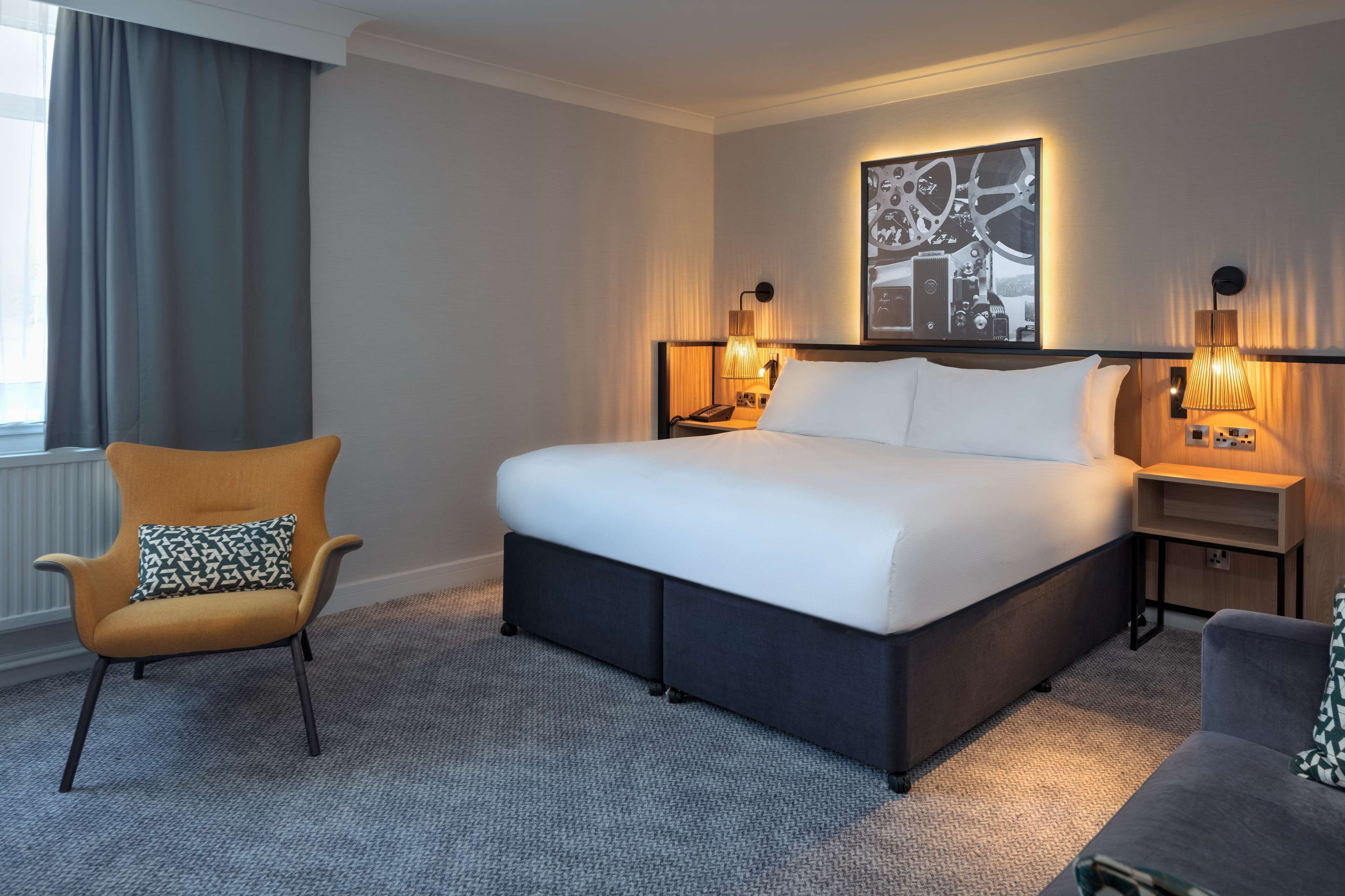 Una habitación de hotel con una cama grande, una silla, una mesita de noche y una lámpara de pared.