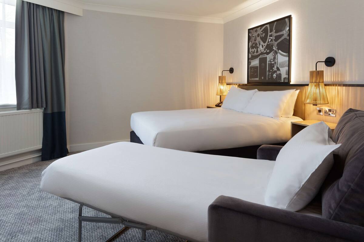 A hotel room with two beds, a sofa, a wall-mounted lamp, and a framed picture.