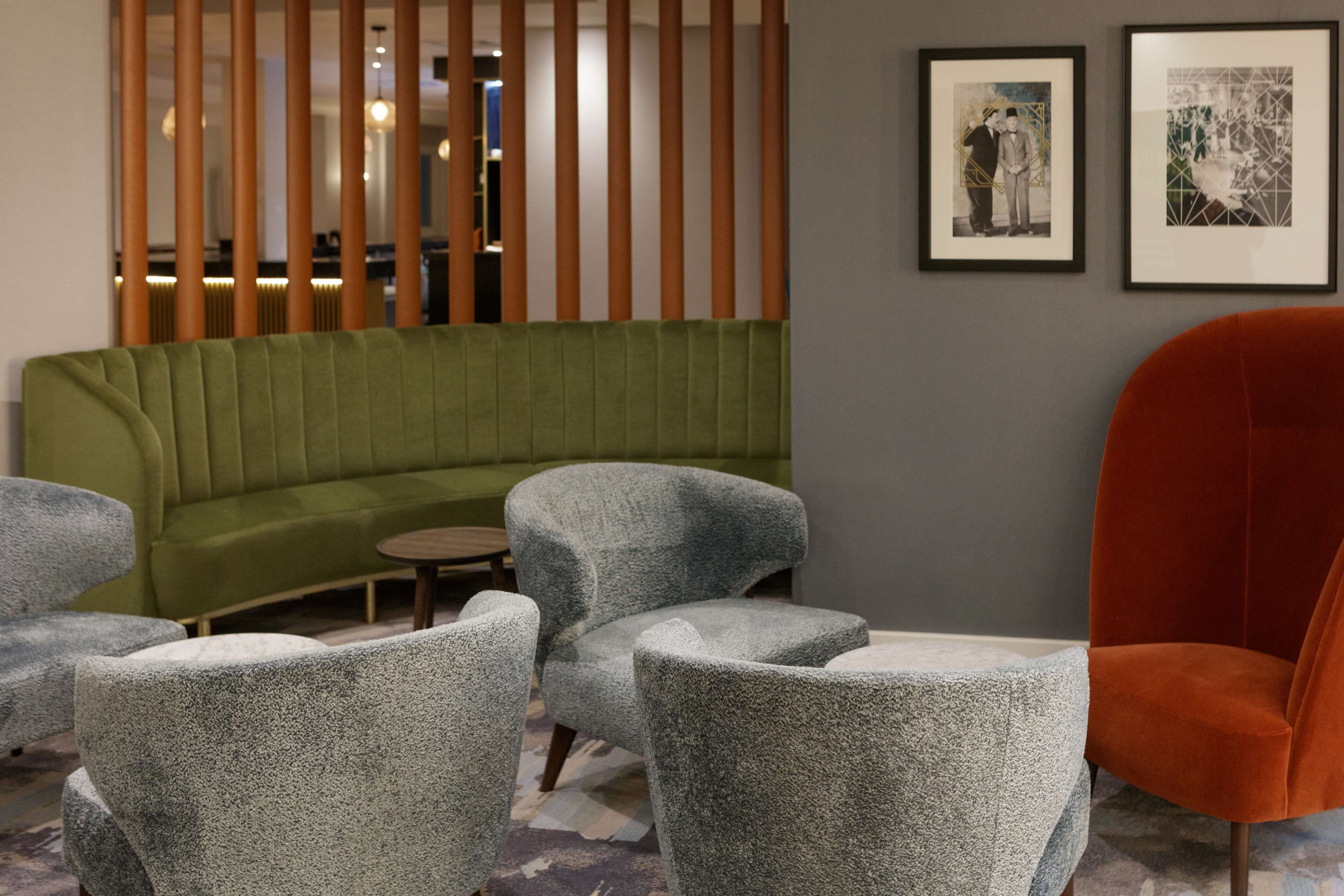 A modern lounge area with a green sofa, grey armchairs, and a red chair. There are framed pictures on the wall.