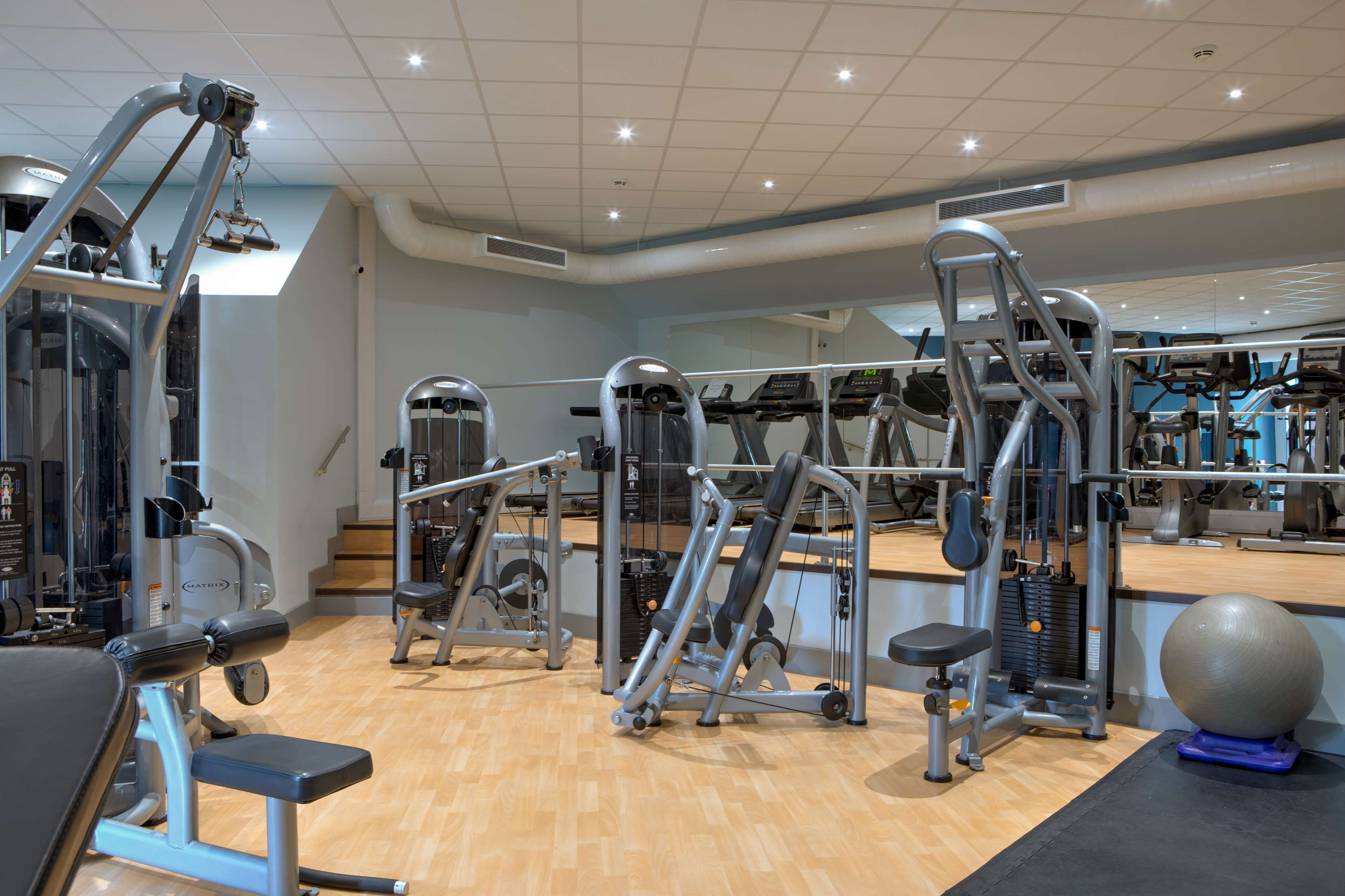 Une salle de sport équipée de divers appareils d’exercice, dotée d’un mur recouvert de miroirs et d’un plancher en bois.