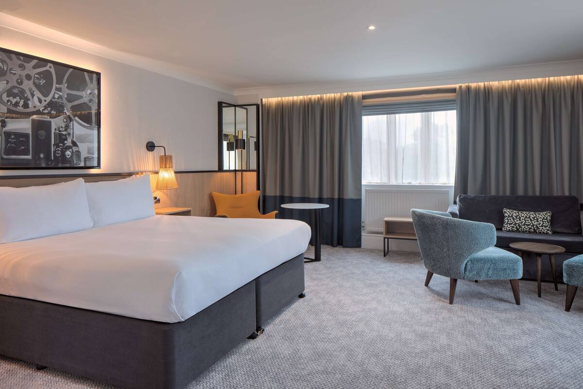 A modern hotel room with a large bed, a sofa, a chair, a small table, and a large window with curtains.