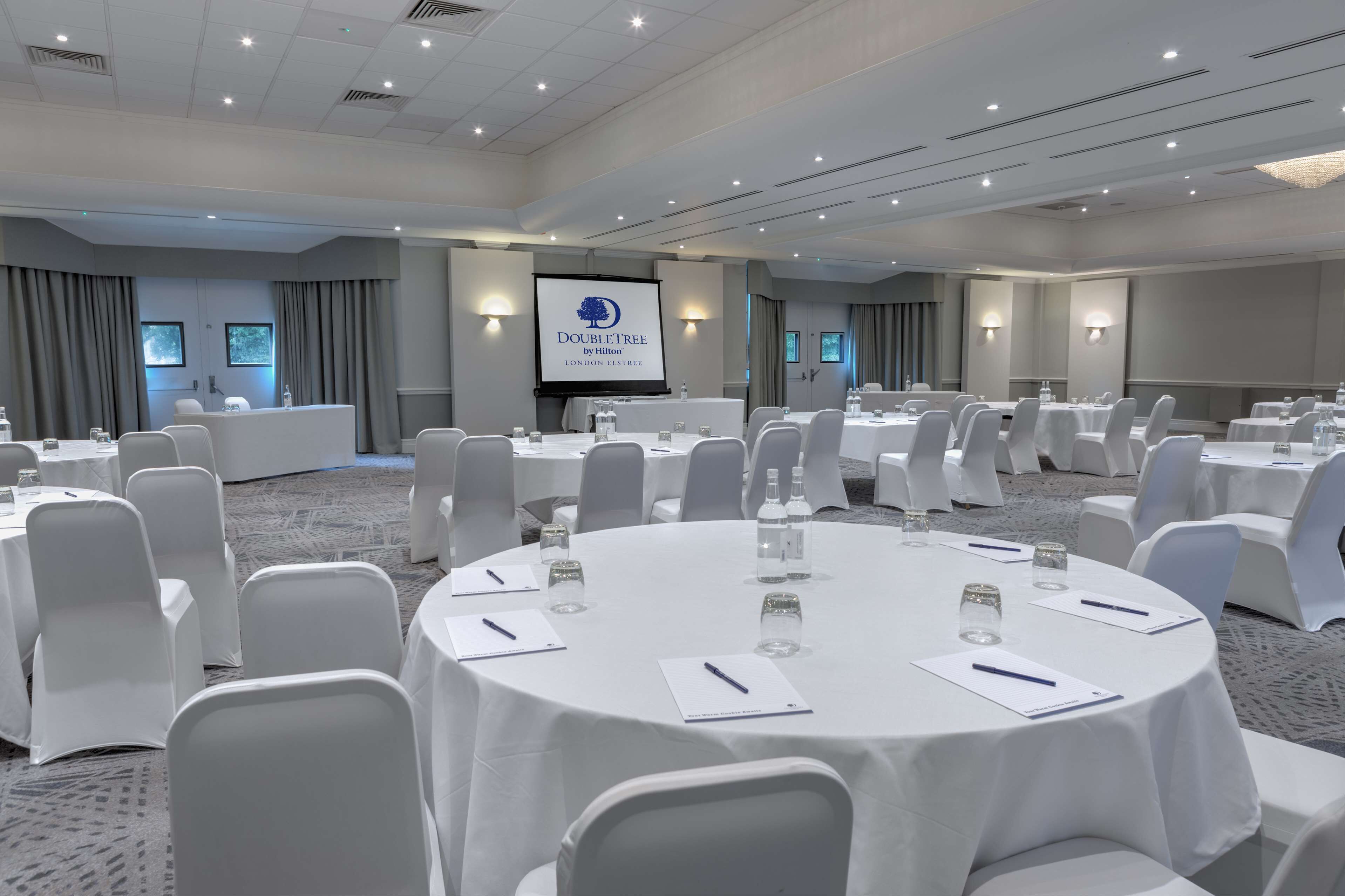 Ein Konferenzraum mit runden Tischen, weißen Stühlen und einer Projektionsleinwand mit dem DoubleTree by Hilton Logo.