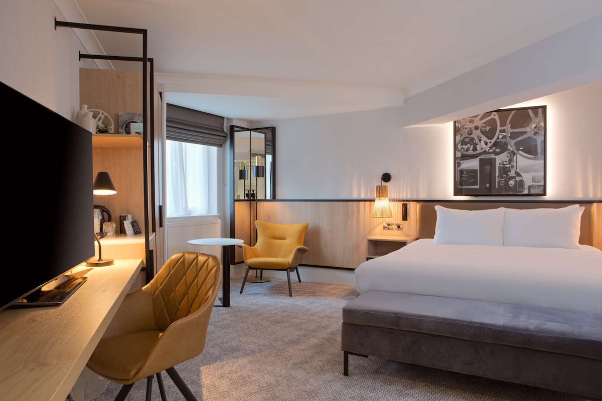 A hotel room with a bed, a desk with a lamp, a chair, a TV, and a large painting on the wall.