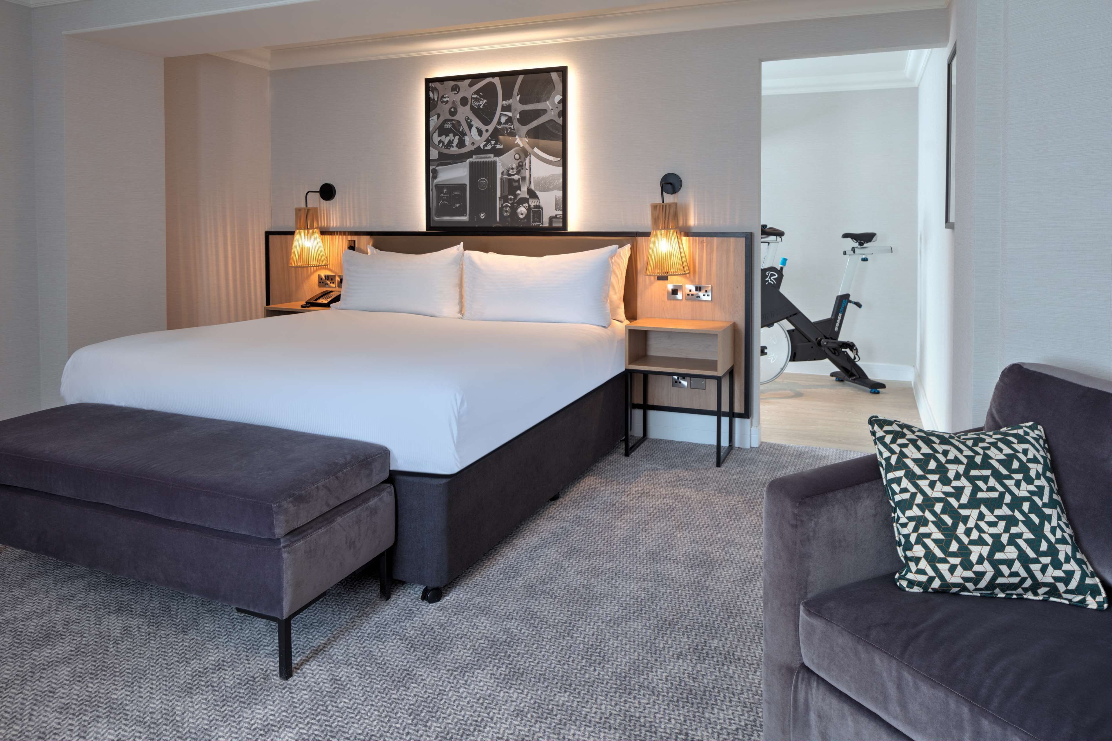 A modern hotel room with a large bed, a sitting area with a sofa and ottoman, a desk with a chair, and a wall-mounted exercise bike.