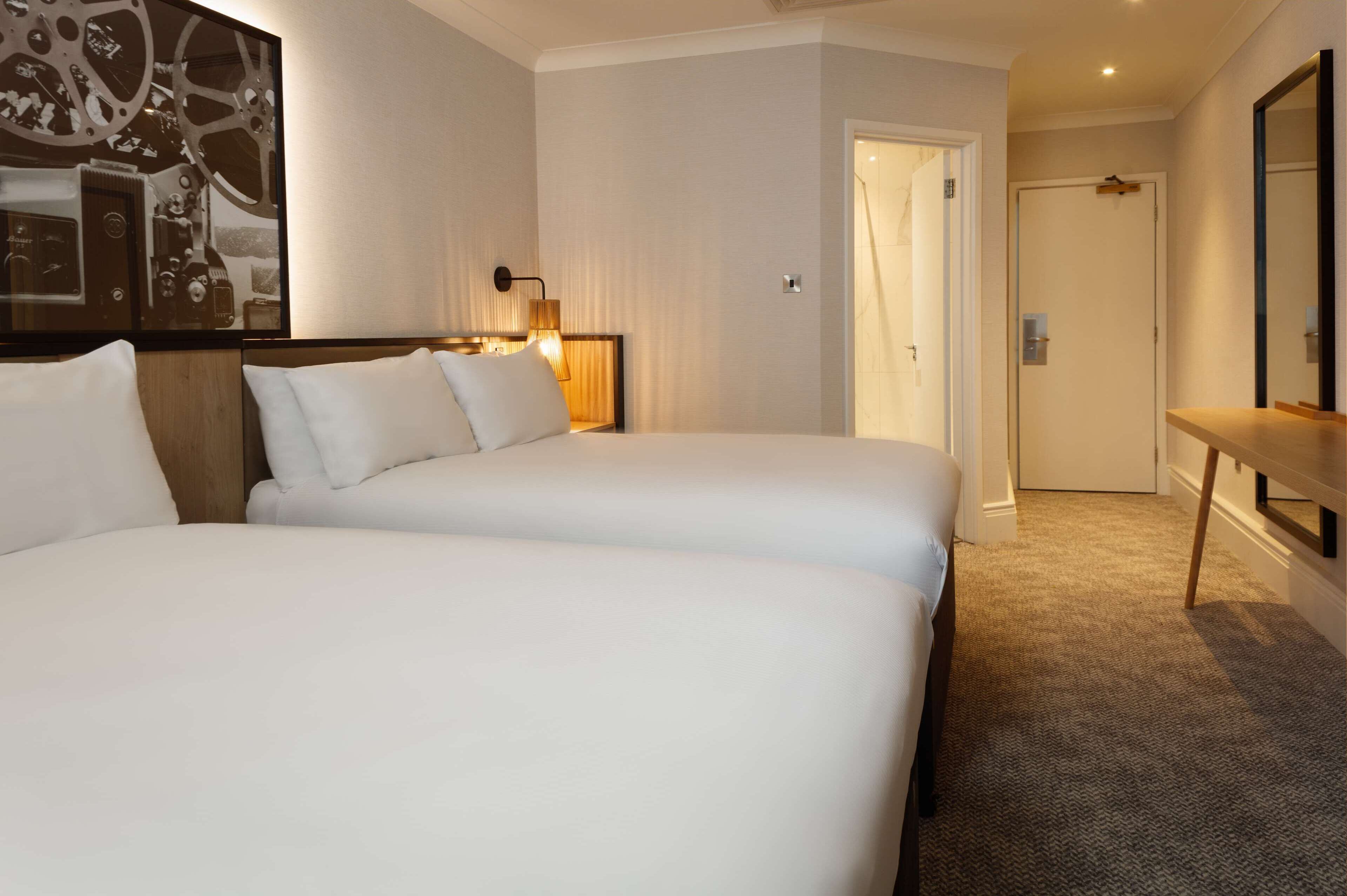 A hotel room with two beds, a wooden headboard, a wall-mounted clock, and a door leading to a bathroom.